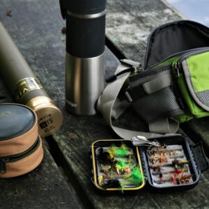 A detailed display of fishing equipment including fly box, backpack, and thermos, set outdoors.